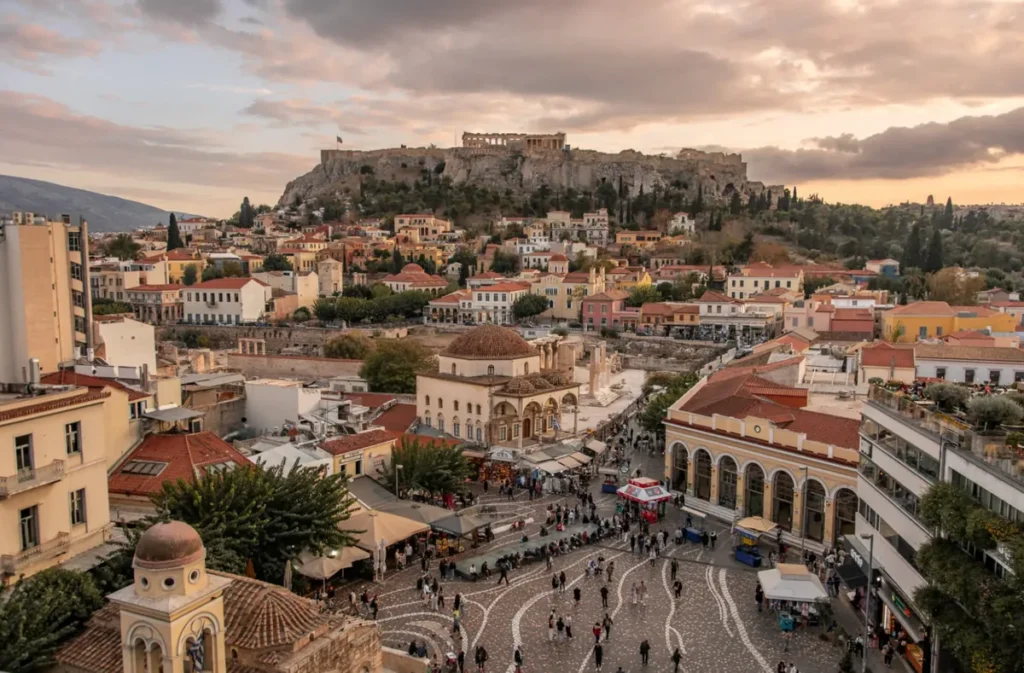 Vista panorâmica do centro histórico de Atenas, capital da Grécia, com a imponente Acrópole e o Partenon dominando a paisagem no alto de uma colina rochosa. Em primeiro plano, a movimentada Praça Monastiraki apresenta seu característico calçamento decorativo, visitantes, estabelecimentos comerciais e a histórica Mesquita Tzistarakis, reconhecida por sua cúpula. O cenário reúne construções neoclássicas, edifícios tradicionais e ruas comerciais que representam a diversidade arquitetônica da capital grega. A iluminação suave do entardecer e o céu com nuvens rosadas destacam a atmosfera histórica e cultural de Atenas.