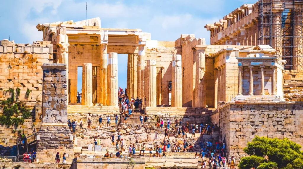 Vista detalhada dos Propileus, a entrada monumental da Acrópole de Atenas, um dos conjuntos arqueológicos mais importantes da Grécia Antiga. A fotografia mostra as grandes colunas e estruturas de mármore da entrada histórica, construída no século V a.C., além das escadarias de acesso percorridas por visitantes. À direita, destaca-se o pequeno Templo de Atena Nice, dedicado à deusa da vitória, construído em estilo jônico. A composição evidencia os elementos da arquitetura clássica grega, as pedras milenares e os trabalhos de conservação do patrimônio arqueológico. Sob o céu azul, o cenário representa a riqueza histórica e cultural de Atenas, um dos destinos mais importantes para conhecer a civilização grega antiga.