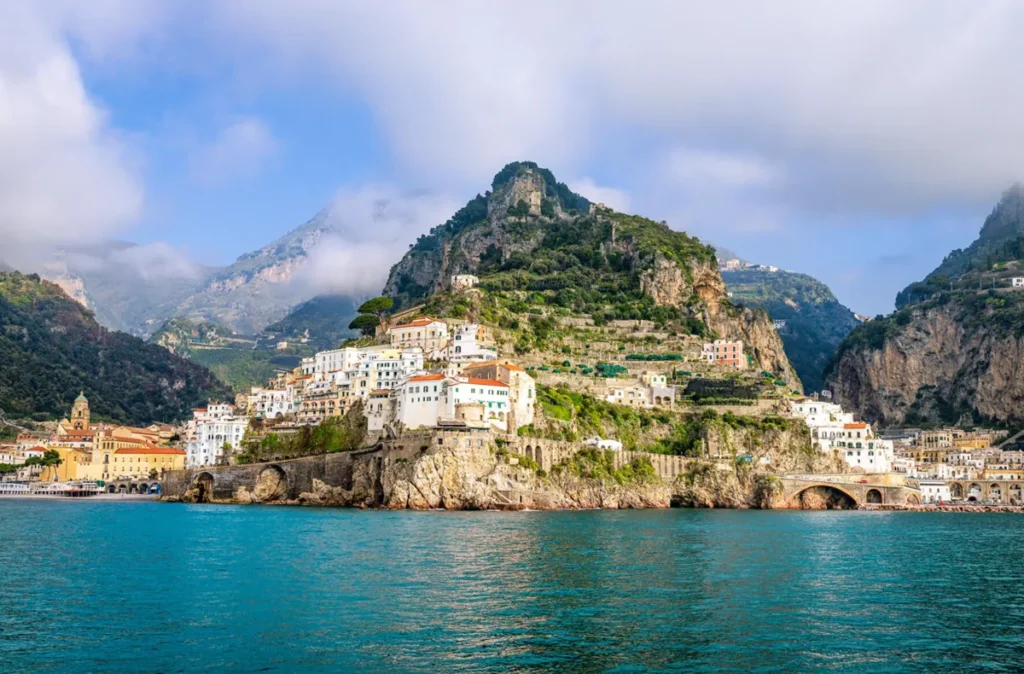Vista panorâmica da cidade de Amalfi, um dos destinos turísticos mais emblemáticos da Costa Amalfitana, na região da Campânia, sul da Itália. A fotografia mostra o centro histórico de Amalfi, com edifícios de fachadas claras e telhados de terracota distribuídos pelas encostas montanhosas que se elevam diretamente do mar Tirreno. A paisagem reúne falésias rochosas, terraços verdes, construções mediterrâneas e antigas estruturas de pedra junto à costa. Ao fundo, as montanhas parcialmente encobertas por nuvens destacam a geografia acidentada desse trecho do litoral italiano, reconhecido pela UNESCO como Patrimônio Mundial.