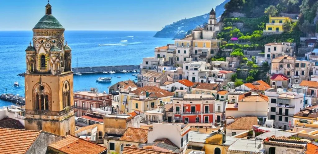 Panorama do centro histórico de Amalfi, na Costa Amalfitana, destacando o campanário da Catedral de Santo André, também conhecida como Duomo di Amalfi, um dos principais monumentos religiosos da cidade. A torre apresenta elementos arquitetônicos medievais e decoração policromada característica da influência árabe-normanda no sul da Itália. Ao redor, edifícios históricos de fachadas claras e telhados de terracota formam um conjunto urbano compacto entre as montanhas e o mar Tirreno. A fotografia também revela o pequeno porto de Amalfi, com barcos ancorados, um quebra-mar e águas azuis que se estendem até o horizonte.