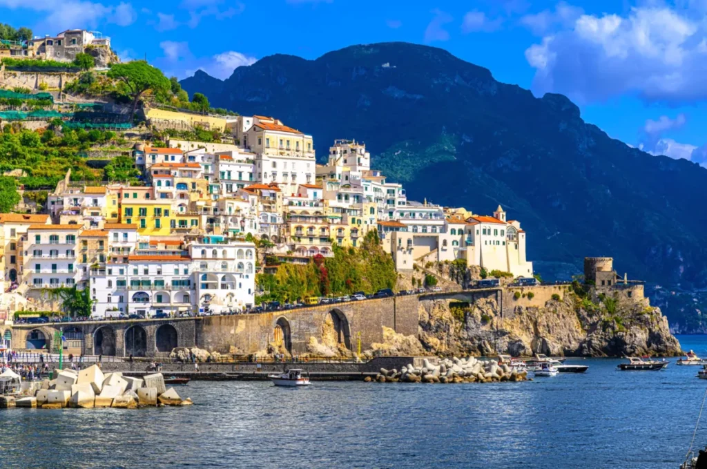 A cidade de Amalfi apresenta uma das paisagens mais características da Costa Amalfitana, combinando arquitetura mediterrânea, montanhas íngremes e o azul intenso do mar Tirreno. A fotografia destaca as casas históricas em tons de branco, amarelo e bege, construídas em diferentes níveis sobre as encostas rochosas, além de hotéis com varandas, terraços ajardinados e uma antiga torre de defesa costeira. Na parte inferior, o porto de Amalfi reúne pequenas embarcações, um quebra-mar e estruturas de pedra que acompanham o litoral. A composição evidencia a integração entre a cidade histórica, a paisagem natural e a tradição marítima do sul da Itália.
