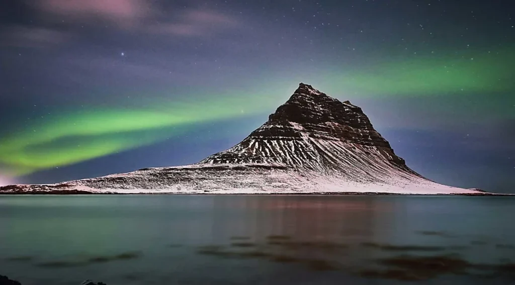 Monte Kirkjufell, na península de Snæfellsnes, no oeste da Islândia, iluminado pelas luzes verdes da aurora boreal durante uma noite de inverno. Conhecida por seu formato cônico característico, essa montanha é um dos lugares mais fotografados do país e um cenário icônico para observar e fotografar a aurora boreal.