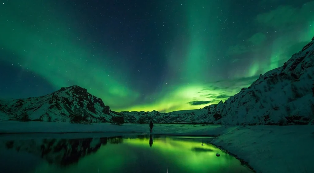 Aurora boreal na Islândia, com intensas luzes verdes dançando sobre montanhas nevadas e refletindo nas águas tranquilas de um lago. O fenômeno natural, também conhecido como Northern Lights, é uma das principais atrações do inverno islandês, especialmente durante noites escuras e com céu limpo, quando a atividade geomagnética favorece sua observação.