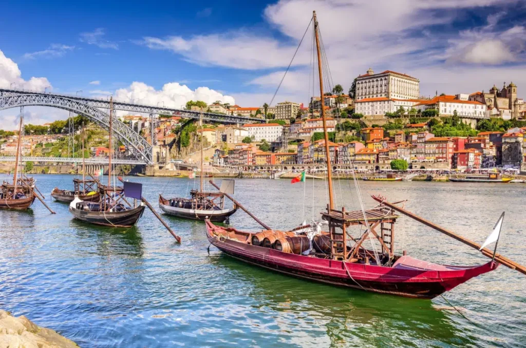 Os tradicionais barcos rabelos são símbolos da história do vinho do Porto e do transporte fluvial no rio Douro, em Portugal. A fotografia destaca diversas embarcações de madeira, incluindo um barco de casco vermelho carregado com barris, diante da Ponte Dom Luís I, uma das estruturas metálicas mais famosas do Porto. Ao fundo, o casario histórico da Ribeira se espalha pelas encostas, acompanhado de igrejas, edifícios monumentais e fachadas coloridas. A cena combina patrimônio arquitetônico, tradição vinícola e a paisagem do rio Douro, que conecta o Porto a Vila Nova de Gaia.