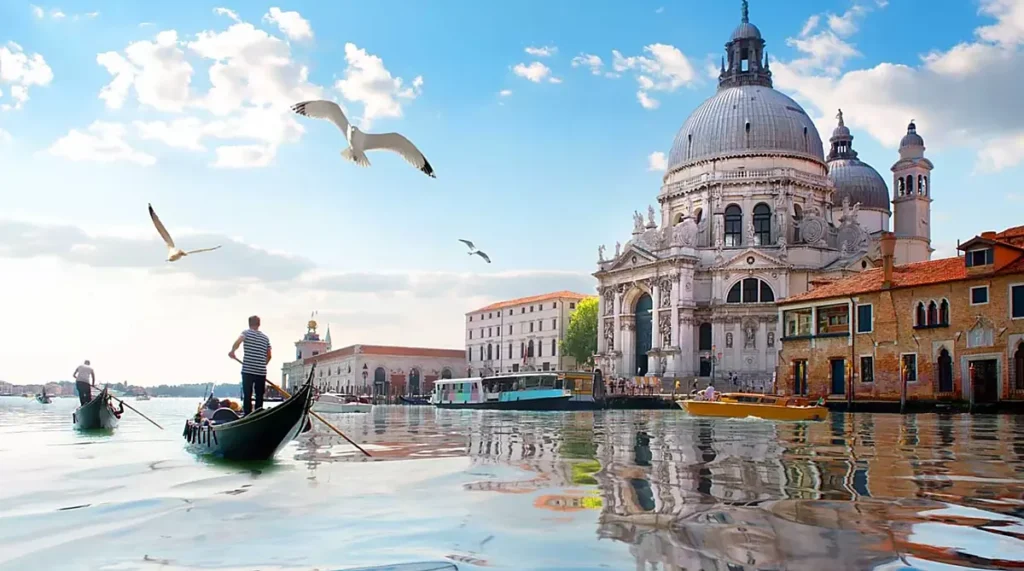 O Grande Canal de Veneza oferece uma das paisagens mais famosas da Itália, combinando gôndolas tradicionais, embarcações históricas e monumentos arquitetônicos às margens da água. Nesta fotografia, gondoleiros conduzem suas embarcações diante da Basílica de Santa Maria della Salute, igreja barroca do século XVII conhecida por suas grandes cúpulas e fachada ornamentada. As gaivotas sobrevoando o canal e os reflexos dos edifícios na água completam a paisagem veneziana, um dos cenários mais procurados por turistas que visitam Veneza.