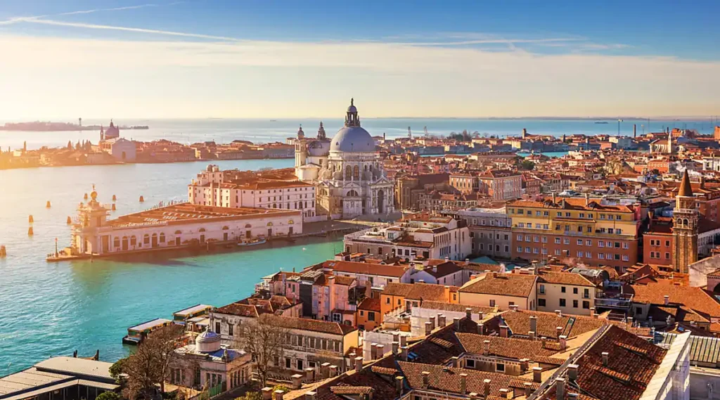 A Basílica de Santa Maria della Salute é um dos principais monumentos religiosos e arquitetônicos de Veneza, localizada próxima à entrada do Grande Canal, no bairro de Dorsoduro. A fotografia panorâmica revela sua monumental cúpula barroca, a Punta della Dogana, as águas da lagoa veneziana e os tradicionais edifícios de telhados avermelhados que caracterizam o centro histórico da cidade. Construída no século XVII, a igreja é um dos símbolos mais reconhecidos da paisagem de Veneza e um dos pontos turísticos mais fotografados da Itália.