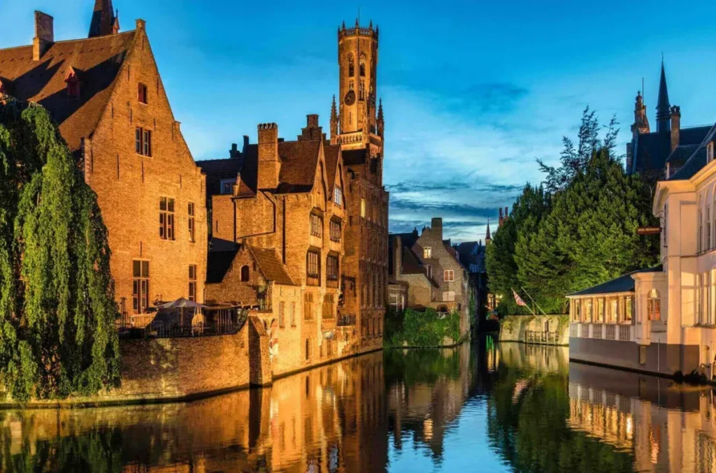 Vista do canal de Rozenhoedkaai, um dos cenários mais emblemáticos de Bruges, na Bélgica, durante o entardecer. A fotografia apresenta construções históricas de tijolos refletidas nas águas tranquilas do canal, enquanto a imponente Torre Belfort se destaca ao fundo contra o céu azul. A iluminação dourada dos edifícios cria uma atmosfera romântica, valorizando os detalhes da arquitetura medieval e a beleza dos canais que atravessam a cidade. Localizada próxima à Praça do Mercado (Markt), a Torre Belfort é um dos principais símbolos de Bruges e oferece uma vista panorâmica do centro histórico. A região de Rozenhoedkaai é especialmente procurada por visitantes e fotógrafos por reunir alguns dos elementos mais característicos da paisagem urbana de Bruges.