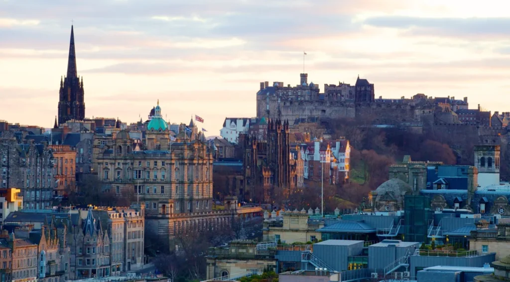Vista panorâmica do centro histórico de Edimburgo, na Escócia, com destaque para o imponente Castelo de Edimburgo, localizado sobre a formação rochosa vulcânica conhecida como Castle Rock. A fortaleza domina a paisagem urbana e está cercada por edifícios históricos, igrejas de torres pontiagudas e construções de arquitetura georgiana e vitoriana. A fotografia captura a cidade sob a iluminação suave do entardecer, revelando o contraste entre as construções de pedra e o céu em tons claros. O Castelo de Edimburgo é um dos monumentos mais emblemáticos da Escócia e uma das principais atrações turísticas do país, oferecendo vistas privilegiadas da capital escocesa.