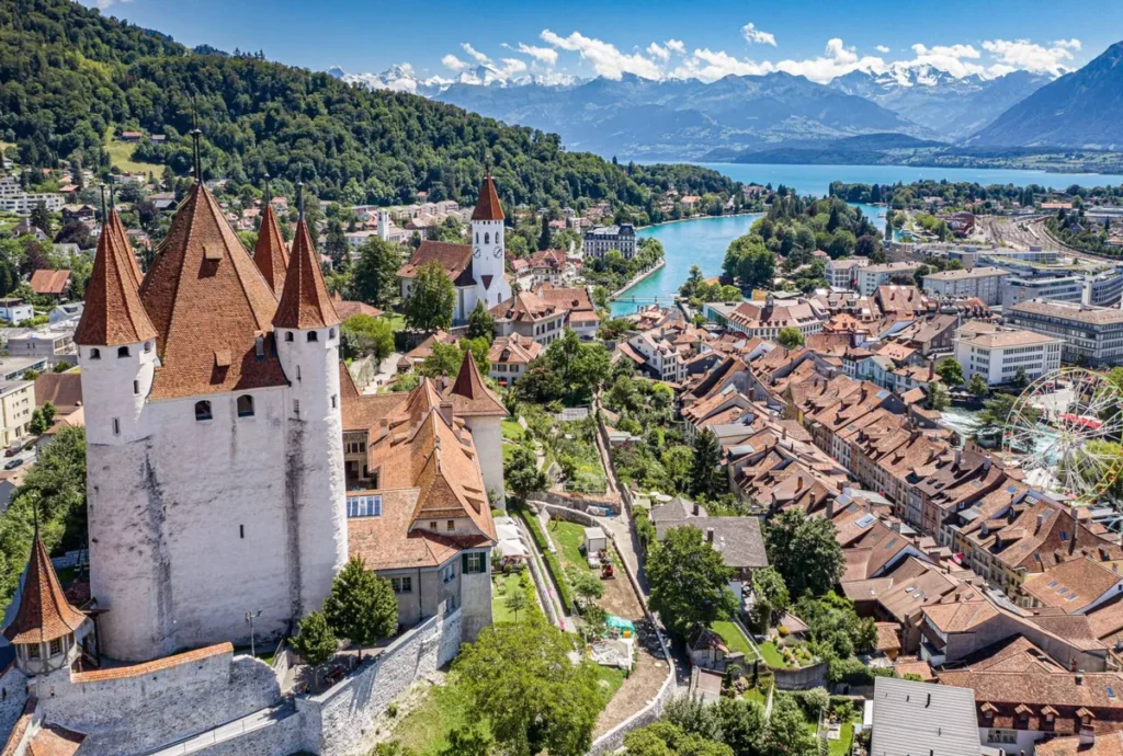 Vista panorâmica da cidade histórica de Thun, situada às margens do Lago Thun, na região do Oberland Bernês, Suíça. Em primeiro plano, destaca-se o imponente Castelo de Thun, uma fortaleza medieval construída no século XII, reconhecida por suas torres e telhados inclinados. Ao redor, o centro histórico apresenta construções tradicionais suíças, igrejas, ruas estreitas e edifícios de telhados avermelhados. Ao fundo, as águas azuladas do Lago Thun e as montanhas dos Alpes Berneses completam uma das paisagens urbanas mais encantadoras da região próxima a Interlaken.