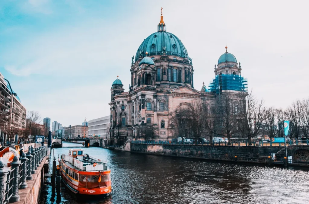 Vista da Catedral de Berlim, conhecida como Berliner Dom, um dos edifícios religiosos e arquitetônicos mais impressionantes da capital alemã. Localizada na Ilha dos Museus, às margens do Rio Spree, a catedral se destaca por sua monumental cúpula esverdeada, fachadas ornamentadas e torres históricas. A fotografia apresenta também uma embarcação turística atracada junto ao rio, elemento característico dos passeios fluviais realizados pelo centro de Berlim. A Berliner Dom é uma das atrações mais visitadas da cidade e integra a paisagem histórica da Ilha dos Museus, região reconhecida como Patrimônio Mundial da UNESCO.