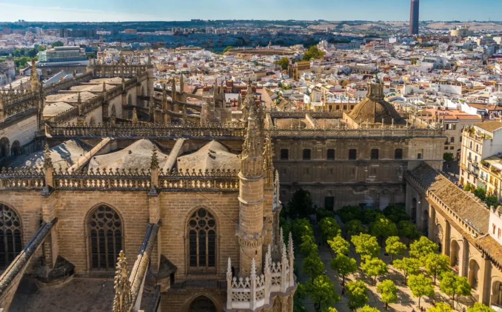 Vista aérea da Catedral de Sevilha, um dos monumentos históricos mais importantes da Andaluzia e uma das maiores catedrais góticas do mundo. A fotografia destaca os detalhes arquitetônicos do edifício, com suas torres ornamentadas, pináculos, vitrais e estruturas monumentais de pedra. Ao lado, aparecem o conjunto arquitetônico histórico e o Patio de los Naranjos, com suas árvores organizadas geometricamente. Ao fundo, a paisagem urbana de Sevilha se estende até o horizonte, revelando os telhados e construções tradicionais da capital andaluza. Reconhecida como Patrimônio Mundial da UNESCO, a Catedral de Sevilha é uma das principais atrações turísticas da Espanha.