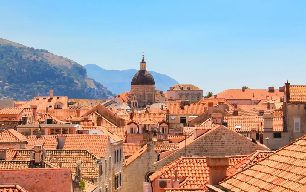 Vista dos tradicionais telhados de terracota do centro histórico de Dubrovnik, na costa da Croácia, com destaque para a cúpula da Catedral da Assunção da Virgem Maria, um dos principais monumentos religiosos da cidade. A fotografia apresenta a arquitetura histórica de Dubrovnik, marcada por construções de pedra calcária, telhados avermelhados e edifícios preservados ao longo dos séculos. Ao fundo, as montanhas que cercam a cidade reforçam a beleza natural da região da Dalmácia. Reconhecida como Patrimônio Mundial da UNESCO, a Cidade Antiga de Dubrovnik é um dos destinos culturais e turísticos mais visitados da Croácia.