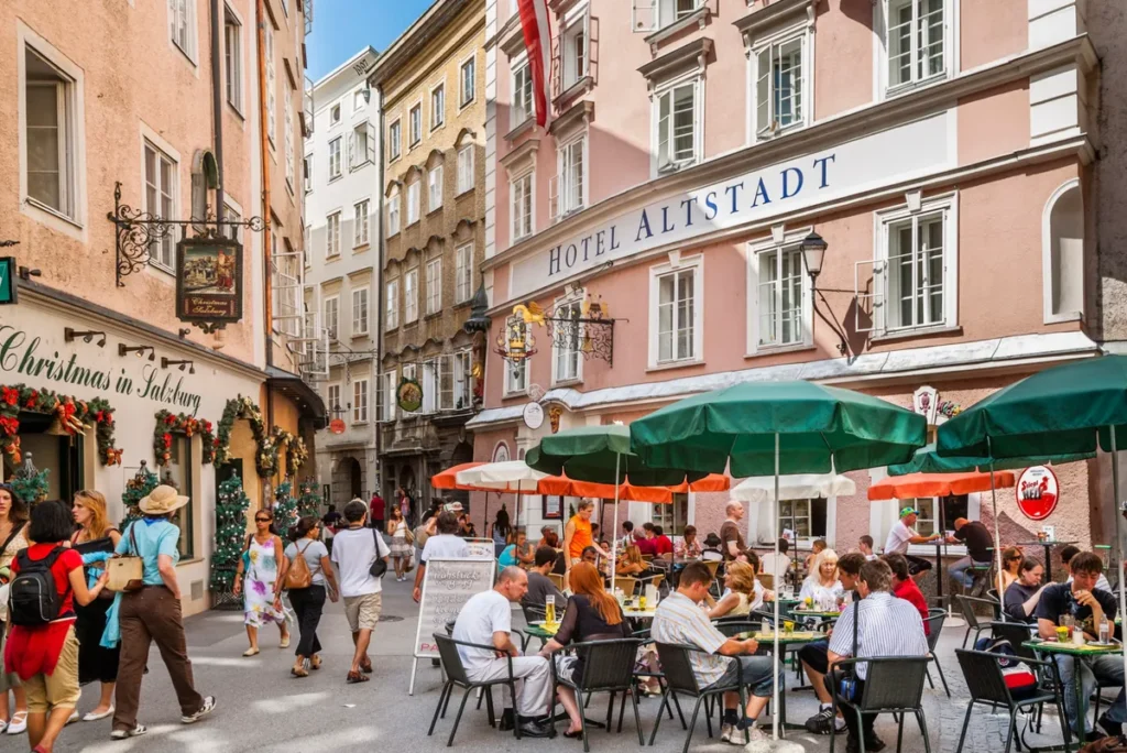 Rua charmosa no centro histórico de Salzburgo, na Áustria, cercada por edifícios antigos de fachadas coloridas, lojas tradicionais e cafés com mesas ao ar livre. A fotografia destaca o movimento de turistas e moradores entre as construções históricas, incluindo o Hotel Altstadt e estabelecimentos que preservam a atmosfera tradicional austríaca. Reconhecido como Patrimônio Mundial pela UNESCO, o centro histórico de Salzburgo encanta por sua arquitetura barroca, ruas estreitas, praças elegantes e pela forte ligação com a vida e a obra do compositor Wolfgang Amadeus Mozart. A região é uma das principais atrações turísticas da cidade e oferece uma combinação de história, cultura, gastronomia e passeios a pé.