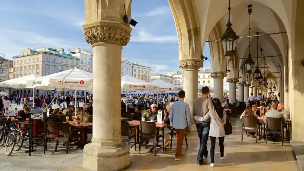 Cafés e restaurantes nas arcadas do Mercado de Tecidos, na Praça do Mercado de Cracóvia. O local combina arquitetura histórica, gastronomia e movimento turístico, oferecendo um ambiente agradável para conhecer o centro histórico da cidade e aproveitar a atmosfera das praças europeias.