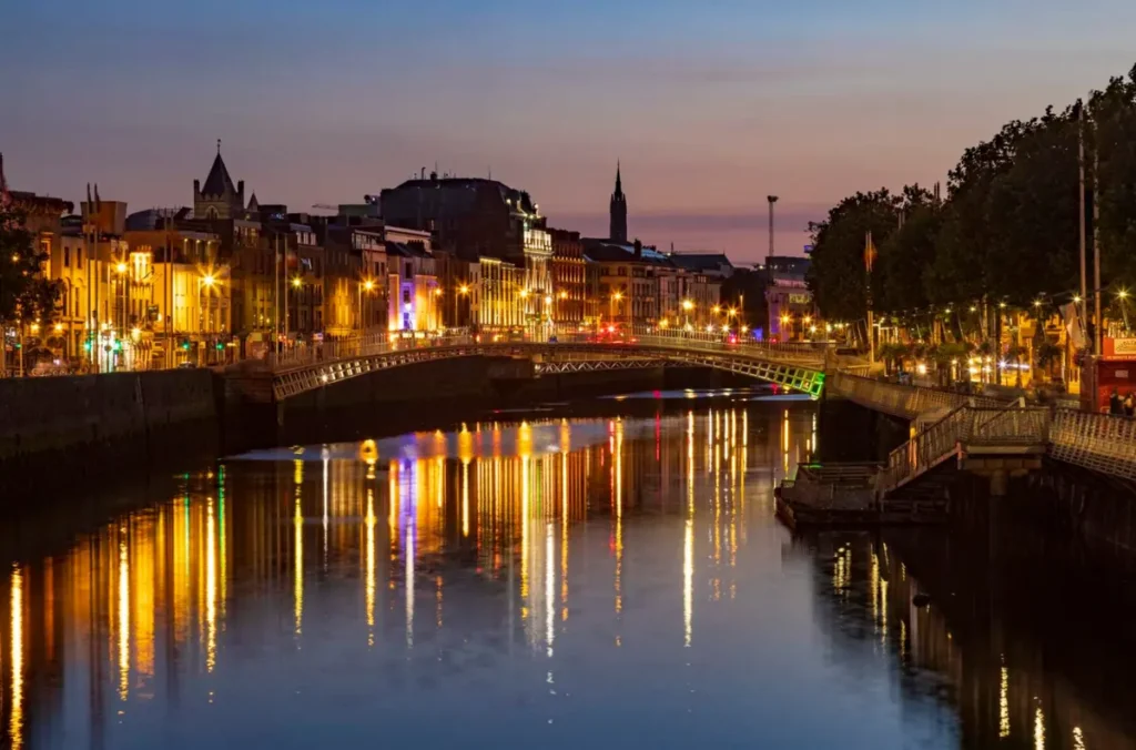 Ha'penny Bridge iluminada ao anoitecer, atravessando o rio Liffey no coração de Dublin. A tradicional ponte de ferro é um dos cartões-postais da capital da Irlanda, especialmente ao entardecer, quando as luzes da cidade se refletem nas águas do rio.