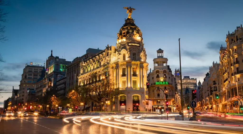 O Edifício Metrópolis é um dos monumentos arquitetônicos mais emblemáticos de Madrid, localizado no encontro da Gran Vía com a Calle de Alcalá, no centro da capital espanhola. A fotografia registra sua fachada monumental iluminada durante a hora azul, destacando a cúpula escura coroada pela escultura dourada da Vitória Alada, os detalhes ornamentais e as colunas clássicas. Ao redor, edifícios históricos, letreiros luminosos e rastros de luz produzidos pelo trânsito revelam a atmosfera urbana de Madrid ao anoitecer.