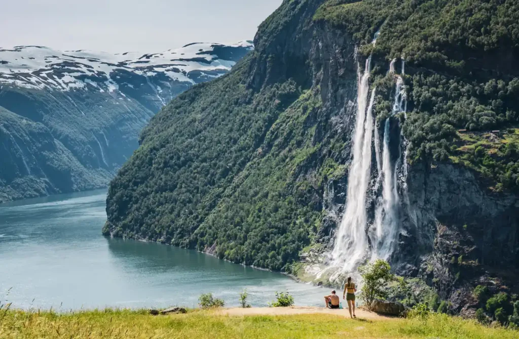 Cachoeiras e montanhas em uma paisagem dos fiordes noruegueses, com quedas-d'água impressionantes, vegetação verde e picos nevados. Cenários como esse são característicos da região de Geirangerfjord, um dos destinos naturais mais famosos da Noruega, reconhecido por seus fiordes profundos e beleza cênica.