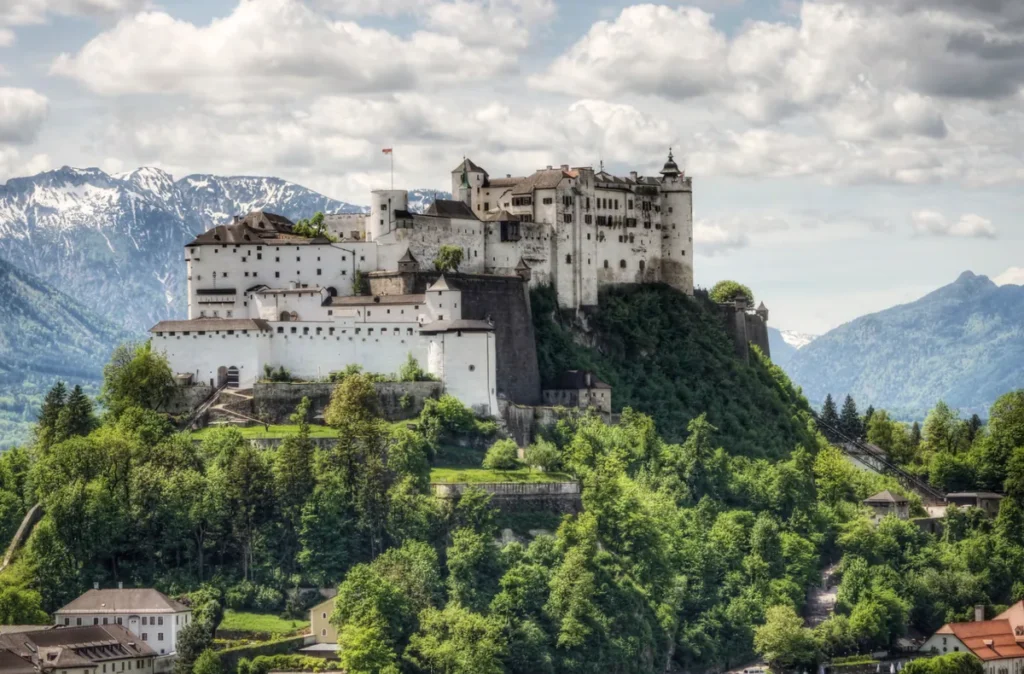Vista panorâmica da Fortaleza de Hohensalzburg, um dos monumentos históricos mais importantes de Salzburgo, na Áustria. Localizada no alto da colina Festungsberg, a fortaleza medieval domina a paisagem da cidade e se destaca por suas muralhas brancas, torres defensivas e grandes construções históricas. Ao fundo, as montanhas dos Alpes austríacos formam um cenário impressionante, enquanto a vegetação verde cobre as encostas ao redor. Sua construção começou em 1077, e o complexo foi ampliado ao longo dos séculos, tornando-se uma das maiores fortalezas medievais preservadas da Europa. Atualmente, é uma das atrações mais visitadas de Salzburgo, oferecendo museus, exposições e vistas panorâmicas da cidade e das montanhas.
