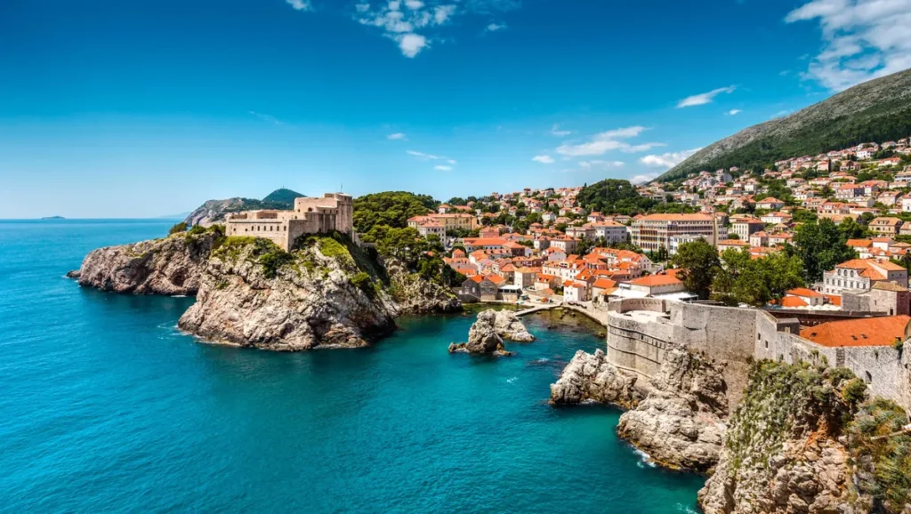Vista panorâmica do litoral de Dubrovnik, na Croácia, com destaque para a imponente Fortaleza Lovrijenac, construída sobre uma formação rochosa às margens do Mar Adriático. A fotografia revela o contraste entre as águas azul-turquesa, os paredões de pedra e as construções históricas da cidade, caracterizadas por fachadas claras e telhados de terracota. Ao fundo, as encostas cobertas de vegetação e as residências distribuídas pelas colinas completam a paisagem mediterrânea. A Fortaleza Lovrijenac é um dos monumentos mais emblemáticos de Dubrovnik e um importante símbolo de sua história defensiva.