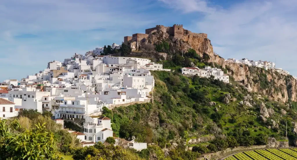 Paisagem de um tradicional pueblo blanco da Andaluzia, com casas brancas espalhadas pela encosta e uma fortaleza histórica sobre formações rochosas. A arquitetura e o cenário natural representam o charme dos vilarejos do sul da Espanha.
