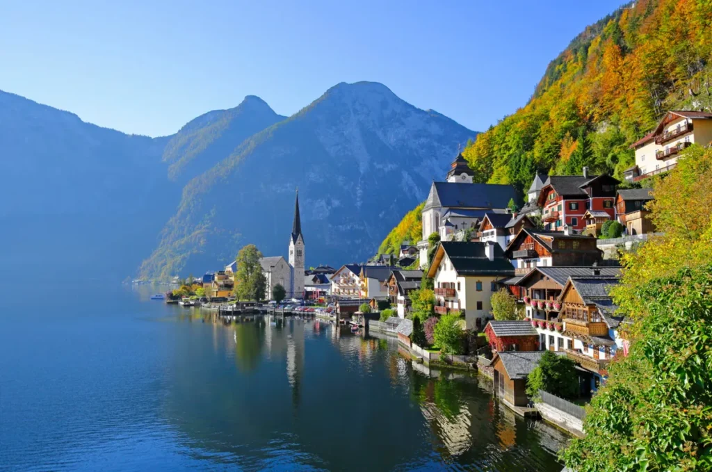 Vista panorâmica de Hallstatt, uma das vilas mais famosas e pitorescas da Áustria, localizada às margens do lago Hallstätter See, na região de Salzkammergut. A fotografia apresenta casas tradicionais de arquitetura alpina, uma igreja com torre pontiaguda e construções distribuídas ao longo da encosta, cercadas por montanhas e florestas com tons de outono. As águas tranquilas do lago refletem parte da paisagem, criando um dos cenários mais fotografados do país. Reconhecida por sua história ligada à mineração de sal e por sua beleza natural, Hallstatt integra uma região classificada como Patrimônio Mundial pela UNESCO. A vila é um destino popular para passeios a partir de Salzburgo, atraindo visitantes interessados em paisagens alpinas, arquitetura histórica e natureza.