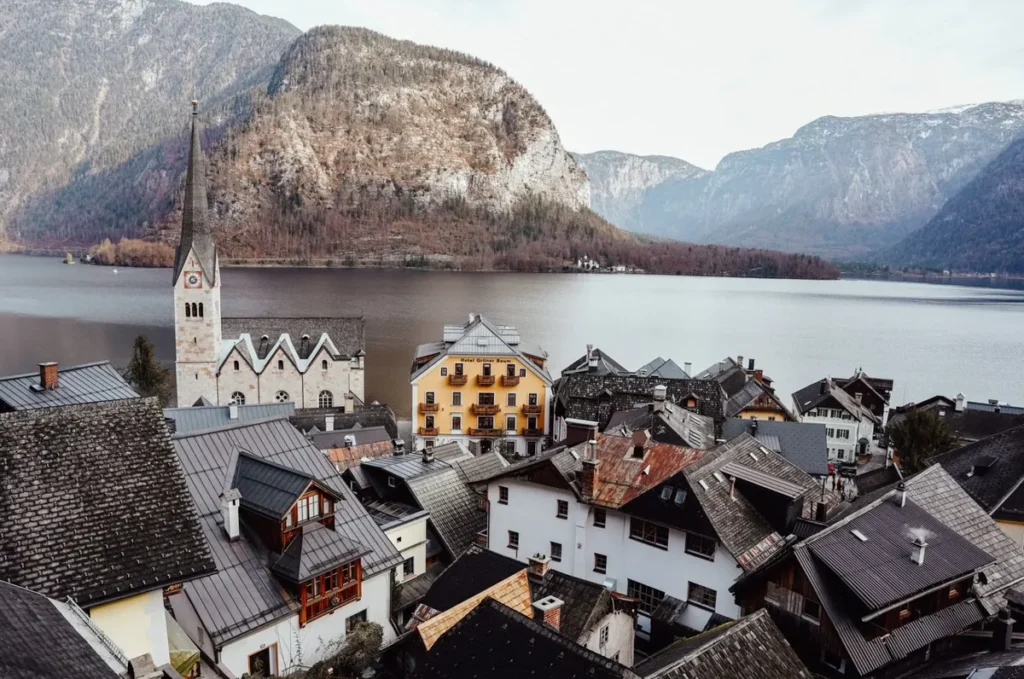 Vista panorâmica da encantadora vila de Hallstatt, localizada às margens do lago Hallstätter See, na região de Salzkammergut, na Áustria. A fotografia destaca a arquitetura tradicional austríaca, com casas históricas de telhados inclinados, construções de madeira e a imponente torre da Igreja Evangélica de Hallstatt, um dos principais cartões-postais da região. Ao fundo, as montanhas dos Alpes austríacos cercam as águas tranquilas do lago, formando uma paisagem de grande beleza natural. Reconhecida por sua história milenar ligada à mineração de sal, Hallstatt integra uma paisagem cultural classificada como Patrimônio Mundial pela UNESCO. A vila é um dos destinos turísticos mais procurados da Áustria e uma opção popular de passeio a partir de Salzburgo, atraindo visitantes interessados em arquitetura histórica, paisagens alpinas e experiências culturais.