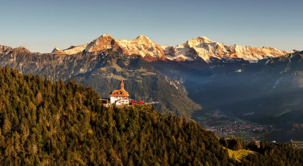 Paisagem panorâmica da região de Interlaken, no coração dos Alpes Berneses, na Suíça. Em primeiro plano, o restaurante panorâmico Harder Kulm destaca-se sobre uma montanha coberta por densas florestas de coníferas. Ao fundo, imponentes picos alpinos cobertos de neve recebem a iluminação dourada do sol, criando um contraste com os vales e as encostas verdes. O Harder Kulm é um dos mirantes mais conhecidos de Interlaken, o