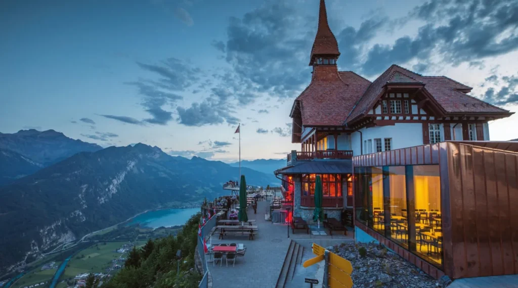 Vista do restaurante panorâmico Harder Kulm ao entardecer, um dos principais mirantes turísticos de Interlaken, na Suíça. O edifício apresenta arquitetura tradicional alpina, com telhados inclinados, detalhes em madeira e grandes janelas iluminadas. O terraço externo oferece vistas impressionantes das montanhas dos Alpes Berneses, do vale de Interlaken e das águas azul-turquesa do Lago Brienz. A iluminação do início da noite cria uma atmosfera acolhedora, tornando o Harder Kulm um destino popular para contemplar o pôr do sol e apreciar a paisagem alpina suíça.