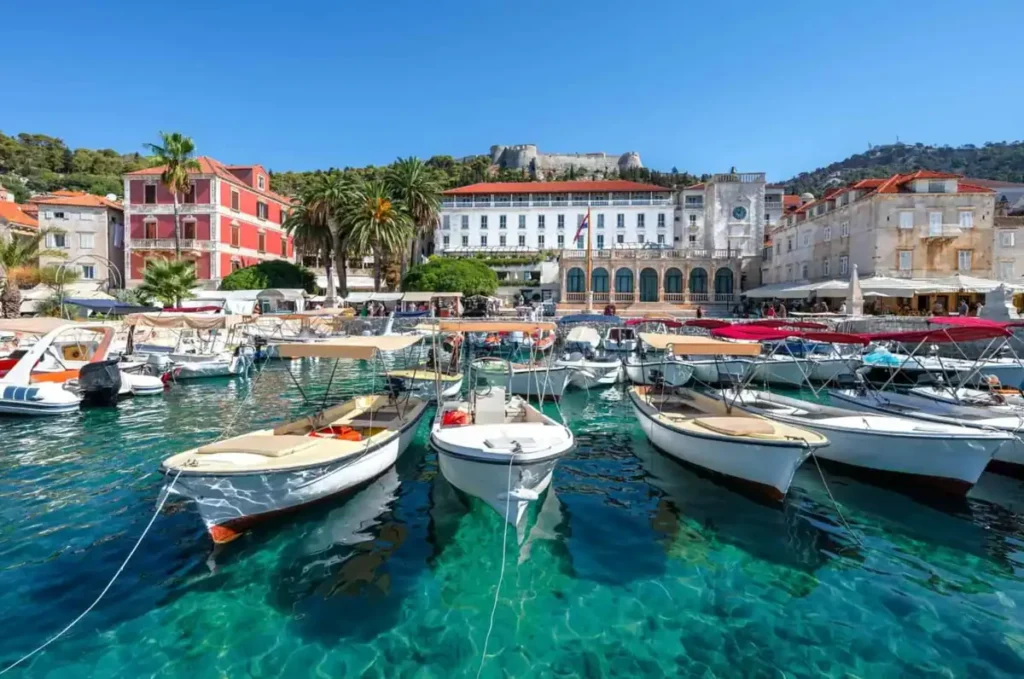 Porto de Hvar, na ilha de Hvar, Croácia, com barcos sobre as águas transparentes do Mar Adriático. Ao fundo, o centro histórico e a Fortaleza Espanhola, também conhecida como Fortica, destacam a arquitetura e as paisagens mediterrâneas de um dos destinos mais famosos da costa da Dalmácia.