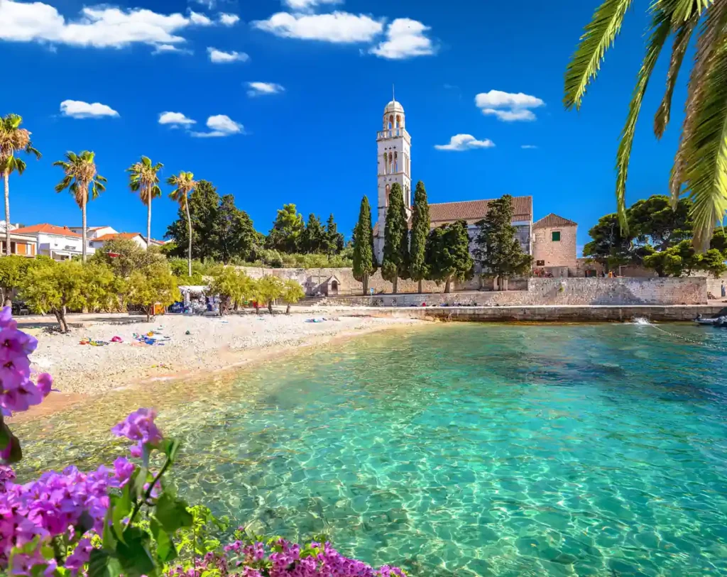 Praia em Hvar, na Croácia, com águas cristalinas do Mar Adriático e vista para o Mosteiro Franciscano. A ilha é conhecida por suas enseadas, praias de pedras claras, vegetação mediterrânea e construções históricas que fazem dela um dos principais destinos turísticos da Dalmácia.