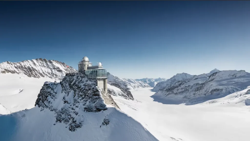 Vista espetacular do Observatório Sphinx, localizado na região de Jungfraujoch, conhecida como Top of Europe, nos Alpes Suíços. A estrutura científica ergue-se sobre uma formação rochosa cercada por montanhas nevadas e pelo extenso glaciar Aletsch, o maior dos Alpes. Situado a aproximadamente 3.571 metros de altitude, o observatório oferece uma das paisagens de alta montanha mais impressionantes da Suíça. A fotografia evidencia o contraste entre a arquitetura moderna, as rochas alpinas e a imensidão branca das geleiras, um dos grandes atrativos turísticos da região de Jungfrau.