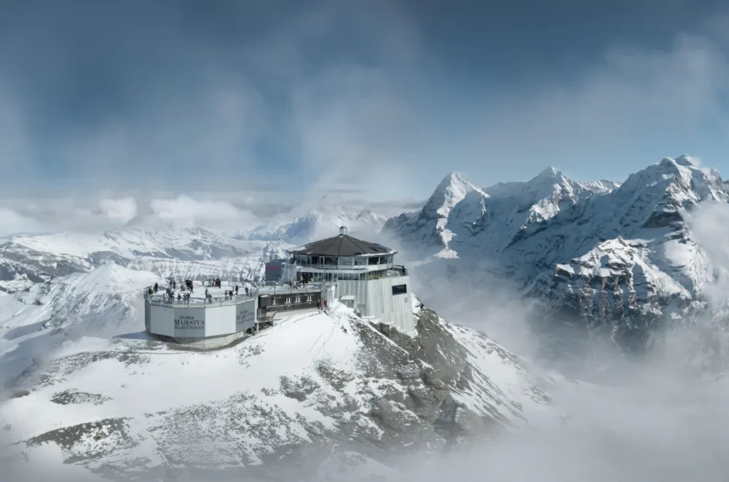 Vista panorâmica do complexo turístico Jungfraujoch – Top of Europe, situado a aproximadamente 3.454 metros de altitude, na região dos Alpes Berneses, Suíça. A fotografia mostra as instalações construídas sobre uma crista montanhosa coberta de neve, incluindo plataformas de observação e áreas destinadas aos visitantes. Ao fundo, os picos nevados dos Alpes Suíços surgem entre nuvens e formações rochosas, criando um cenário de alta montanha. Jungfraujoch é um dos destinos turísticos mais famosos da Suíça, acessível por ferrovia de montanha e conhecido pelas vistas das geleiras e das montanhas da região de Jungfrau.