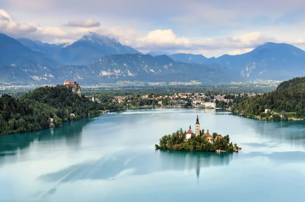 Lago Bled, um dos destinos naturais mais famosos da Eslovênia, com sua ilha e a Igreja da Assunção de Maria. A paisagem inclui o Castelo de Bled sobre um penhasco e as montanhas dos Alpes Julianos, formando um dos cenários mais fotografados do país.