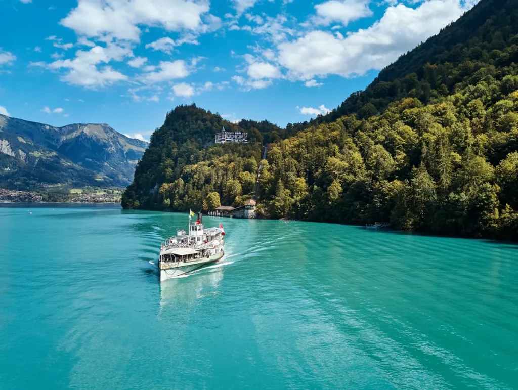 Barco turístico tradicional navegando pelas águas cristalinas de tonalidade azul-turquesa do Lago Brienz, um dos lagos mais bonitos da região de Interlaken, na Suíça. A embarcação atravessa uma paisagem cercada por montanhas cobertas de florestas, pequenas construções alpinas e encostas verdes. Ao fundo, destaca-se a vegetação abundante das montanhas do Oberland Bernês, sob um céu azul com nuvens brancas. O Lago Brienz é conhecido por suas águas de origem glacial e pelos passeios de barco que conectam localidades pitorescas da região, proporcionando vistas panorâmicas dos Alpes Suíços.