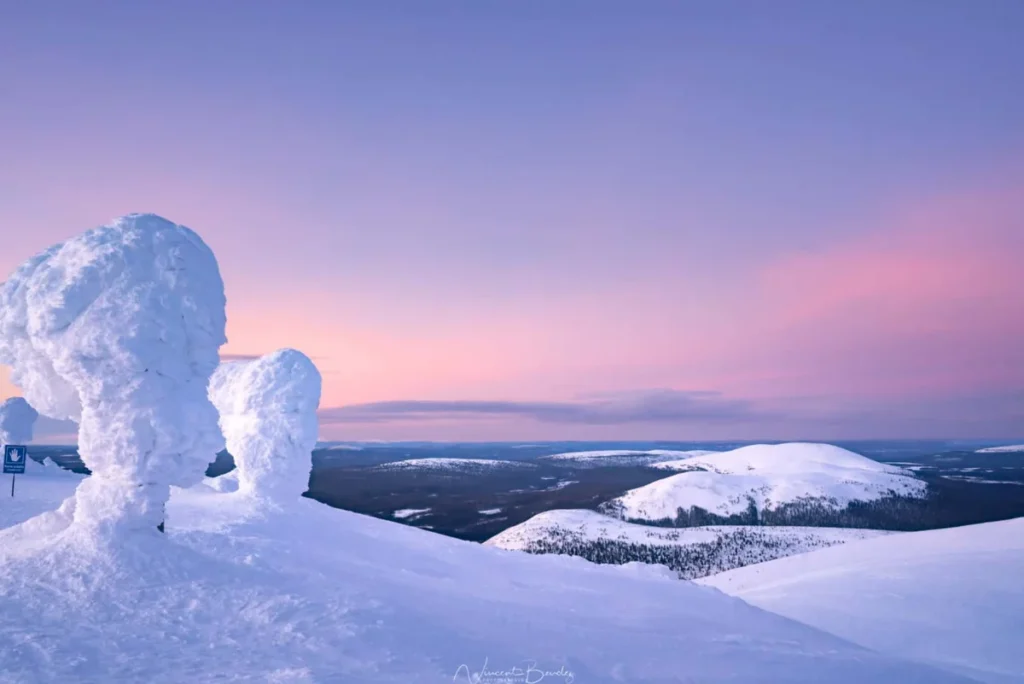 Paisagem de inverno na Lapônia, com árvores congeladas e montanhas cobertas de neve sob um céu colorido pelo crepúsculo ártico. As temperaturas extremamente baixas transformam as florestas da região em esculturas naturais de gelo, criando cenários espetaculares para fotografias e experiências na natureza.