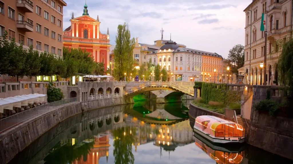 Centro histórico de Ljubljana ao entardecer, com a Ponte Tripla e a Igreja Franciscana da Anunciação iluminadas. O rio Ljubljanica reflete a arquitetura e as luzes da cidade, criando uma das paisagens mais conhecidas da capital da Eslovênia.
