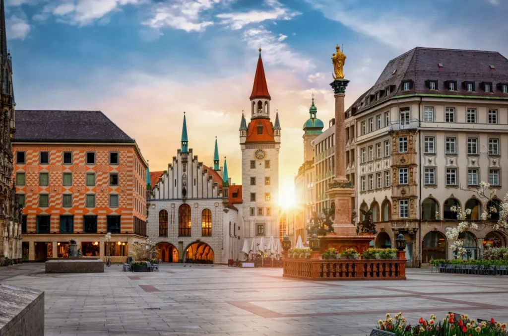 Vista da Marienplatz, no coração de Munique, durante o entardecer, com destaque para a Antiga Prefeitura (Altes Rathaus) e a Coluna de Maria (Mariensäule). A fotografia apresenta a torre histórica da prefeitura, com seu telhado avermelhado e relógio, além da coluna coroada por uma estátua dourada da Virgem Maria, um dos monumentos mais conhecidos da praça. Ao redor, edifícios de diferentes estilos arquitetônicos compõem a paisagem do centro histórico, enquanto a luz do pôr do sol ilumina as fachadas. Considerada o principal ponto de encontro da capital bávara, a Marienplatz reúne importantes atrações turísticas e é um dos locais mais visitados por quem deseja conhecer a história, a cultura e a arquitetura de Munique.