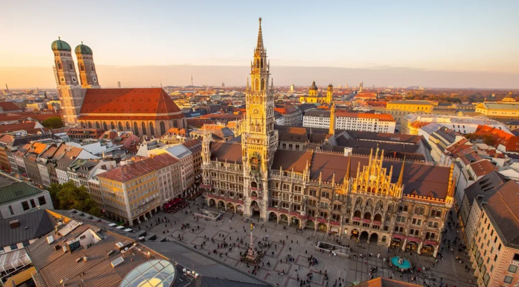 Vista panorâmica da Marienplatz, a praça mais famosa de Munique, na Alemanha, durante o pôr do sol. A fotografia destaca a imponente Nova Prefeitura (Neues Rathaus), um dos principais cartões-postais da cidade, com sua arquitetura neogótica, fachada ornamentada e a torre que abriga o famoso Glockenspiel. À esquerda, aparecem as duas torres da Frauenkirche, a Catedral de Nossa Senhora, reconhecida por suas características cúpulas verdes. Cercada por edifícios históricos, lojas, restaurantes e cafés, a Marienplatz é o coração do centro histórico de Munique e um dos pontos de partida mais populares para explorar as atrações da capital da Baviera.