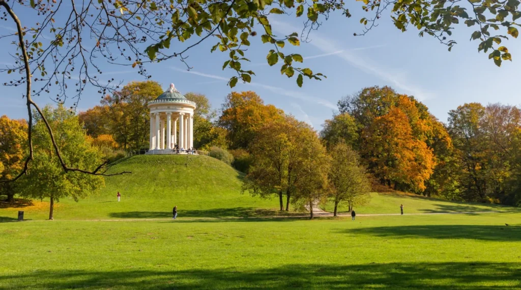 Vista do Monopteros, um elegante templo de inspiração grega localizado no Jardim Inglês (Englischer Garten), um dos parques urbanos mais conhecidos de Munique, na Alemanha. Construído no século XIX, o monumento apresenta colunas brancas e uma cúpula circular, destacando-se sobre uma pequena colina gramada. A fotografia registra a paisagem durante o outono, quando as árvores do parque adquirem tons de amarelo, laranja e dourado, criando um cenário especialmente bonito. O Englischer Garten é um dos principais espaços verdes da capital bávara, muito procurado por moradores e turistas para caminhadas, passeios de bicicleta, piqueniques e momentos de descanso ao ar livre.
