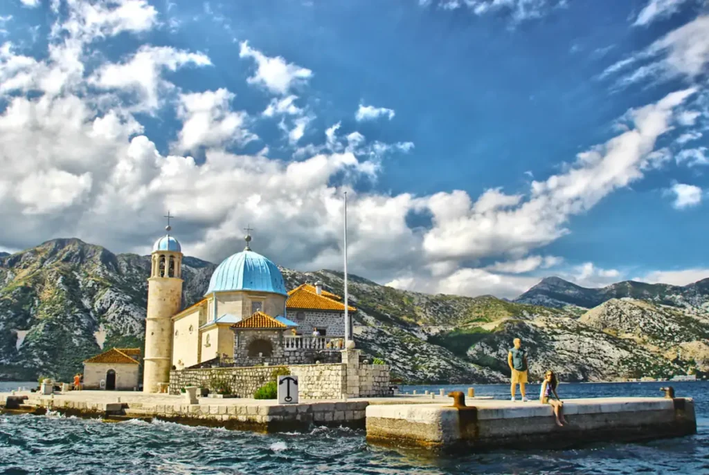 Igreja de Nossa Senhora das Rochas, em uma pequena ilha na Baía de Kotor, Montenegro, cercada por águas azuis e montanhas.