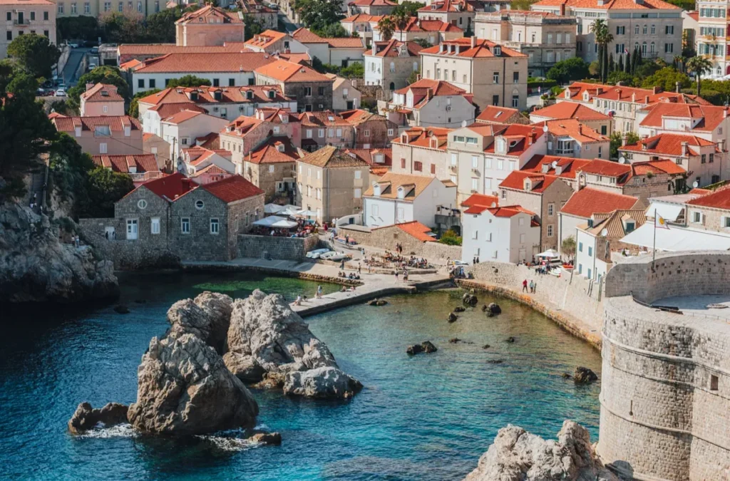 Vista de uma pequena enseada junto às muralhas históricas de Dubrovnik, na Croácia, onde as águas transparentes do Mar Adriático encontram formações rochosas e antigas construções de pedra. A fotografia destaca as imponentes estruturas defensivas da cidade, incluindo uma torre circular de pedra, além das casas tradicionais com telhados de terracota que compõem a paisagem urbana. A região é conhecida pela combinação de arquitetura medieval, praias e cenários naturais mediterrâneos. As muralhas de Dubrovnik estão entre as fortificações históricas mais preservadas da Europa e oferecem vistas panorâmicas do litoral croata.