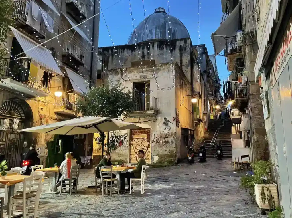 Praça tradicional de Nápoles ao entardecer, com cafés ao ar livre, construções históricas, ruas de pedra e luzes decorativas. A cena retrata o ambiente dos bairros antigos da cidade italiana, onde pequenos restaurantes, escadarias e praças preservam o charme e a vida cotidiana napolitana.