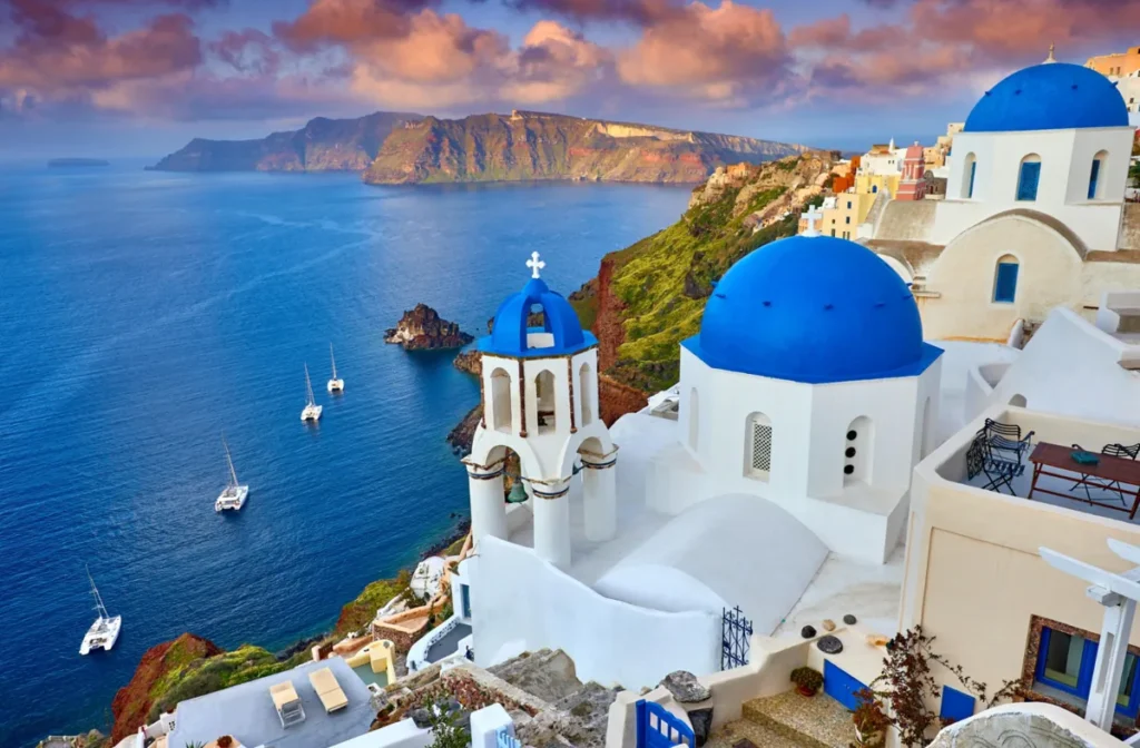 Vista panorâmica da vila de Oia, um dos destinos turísticos mais famosos de Santorini, no arquipélago das Cíclades, Grécia. A fotografia destaca as tradicionais igrejas ortodoxas gregas com cúpulas azuis intensas, fachadas brancas e um campanário coroado por uma cruz, elementos emblemáticos da arquitetura cicládica. As construções estão distribuídas sobre falésias vulcânicas com vista privilegiada para a caldeira de Santorini e as águas profundas do mar Egeu. Pequenos veleiros aparecem ancorados na baía, enquanto as montanhas vulcânicas ao fundo recebem a iluminação suave do entardecer. O céu apresenta nuvens em tons de rosa, laranja e lilás, criando uma das paisagens mais reconhecidas da Grécia.