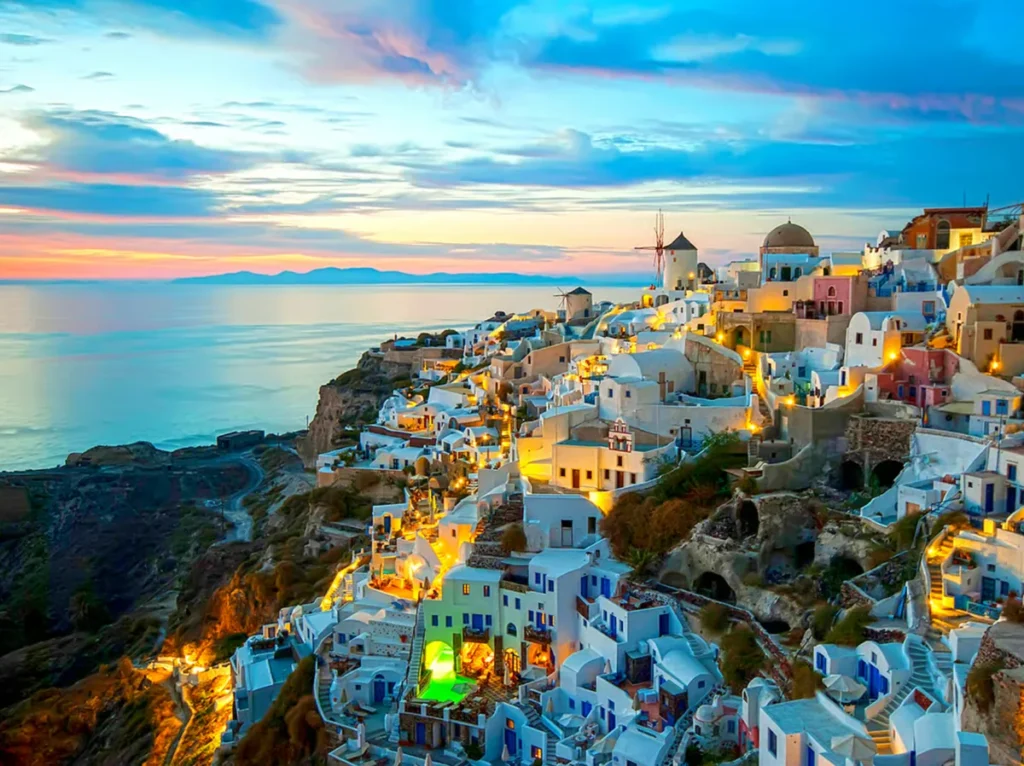 Pôr do sol na vila de Oia, localizada na extremidade norte da ilha de Santorini, na Grécia, um dos lugares mais procurados para apreciar o entardecer no mar Egeu. A fotografia apresenta uma vista elevada das construções tradicionais de arquitetura cicládica, com casas brancas, pequenas igrejas, terraços, escadarias estreitas e moinhos de vento históricos distribuídos ao longo das falésias vulcânicas. As luzes amarelas que iluminam as ruas e os edifícios criam um contraste marcante com o céu em tons de azul, rosa e laranja. Ao fundo, o horizonte do mar Egeu e as silhuetas de ilhas distantes completam a paisagem mediterrânea de Santorini, um dos destinos mais fotografados da Grécia.