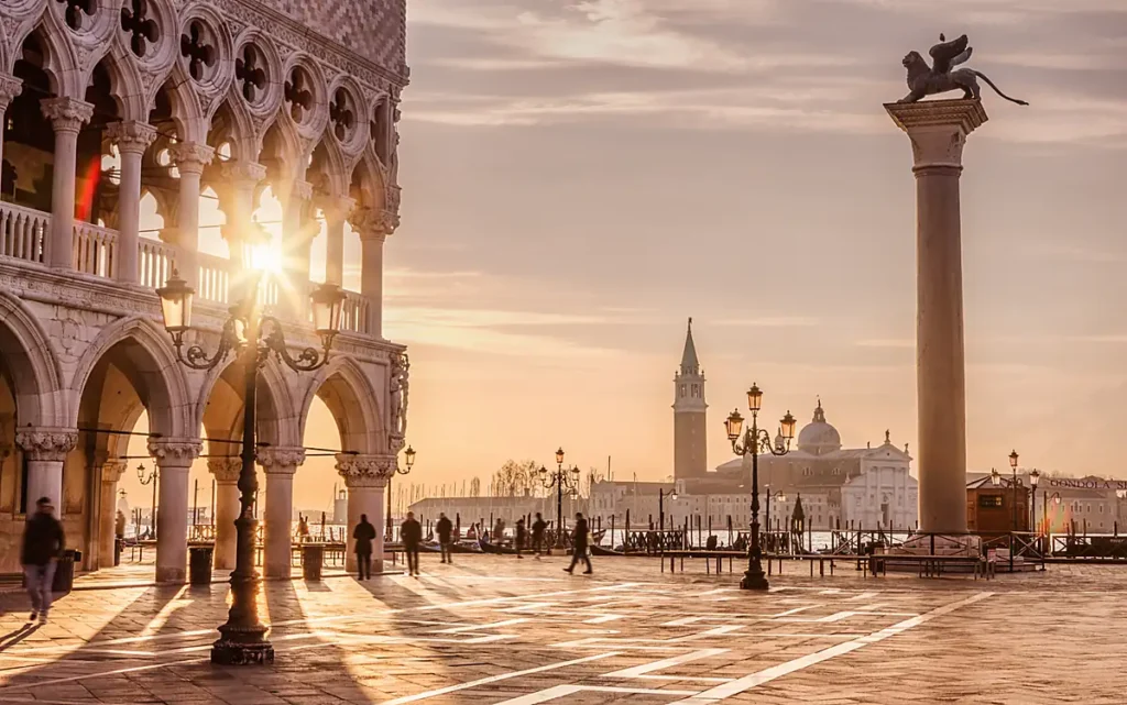A Piazzetta San Marco é um dos cenários históricos mais emblemáticos de Veneza, na Itália. A fotografia destaca as arcadas góticas do Palácio Ducal, antiga sede do poder político da República de Veneza, e a Coluna de São Marcos, coroada pelo tradicional leão alado, símbolo da cidade. Ao fundo, a Igreja de San Giorgio Maggiore aparece do outro lado da lagoa veneziana, enquanto a luz dourada do pôr do sol ilumina a arquitetura histórica e o calçamento da praça.
