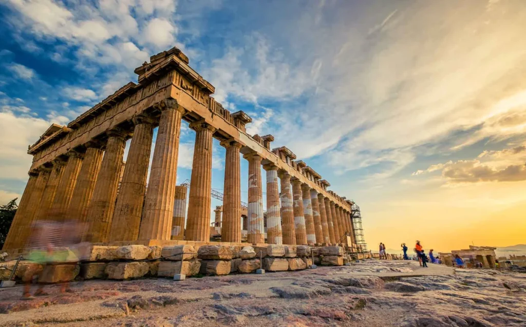 Vista monumental do Partenon, o famoso templo da Grécia Antiga localizado no alto da Acrópole de Atenas. Construído no século V a.C. e dedicado à deusa Atena, o monumento é um dos maiores símbolos da arquitetura clássica grega e do patrimônio histórico europeu. A fotografia apresenta uma perspectiva lateral das imponentes colunas dóricas, destacadas pela luz dourada do entardecer. O céu azul, com nuvens iluminadas em tons amarelos e alaranjados, cria um contraste com a pedra histórica. Visitantes percorrem o sítio arqueológico, um dos principais pontos turísticos de Atenas e Patrimônio Mundial da UNESCO.
