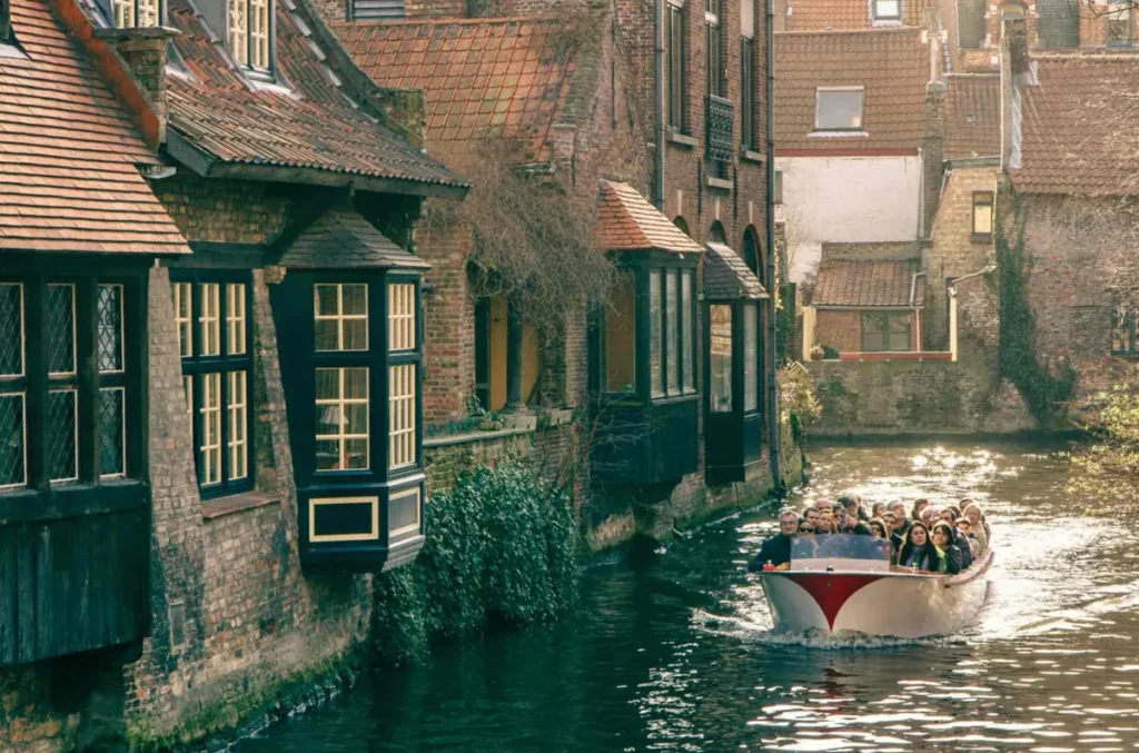 Passeio de barco pelos canais de Bruges, uma das experiências turísticas mais populares da cidade belga. A fotografia mostra uma embarcação transportando visitantes por um canal estreito, cercado por antigas construções de tijolos, janelas ornamentadas e fachadas históricas que parecem surgir diretamente da água. Conhecida como a Veneza do Norte, Bruges possui uma extensa rede de canais que atravessa seu centro histórico e permite admirar a arquitetura medieval de uma perspectiva diferente. Durante o passeio, os visitantes podem observar pontes de pedra, edifícios centenários e paisagens que revelam a atmosfera romântica e o patrimônio arquitetônico da cidade.