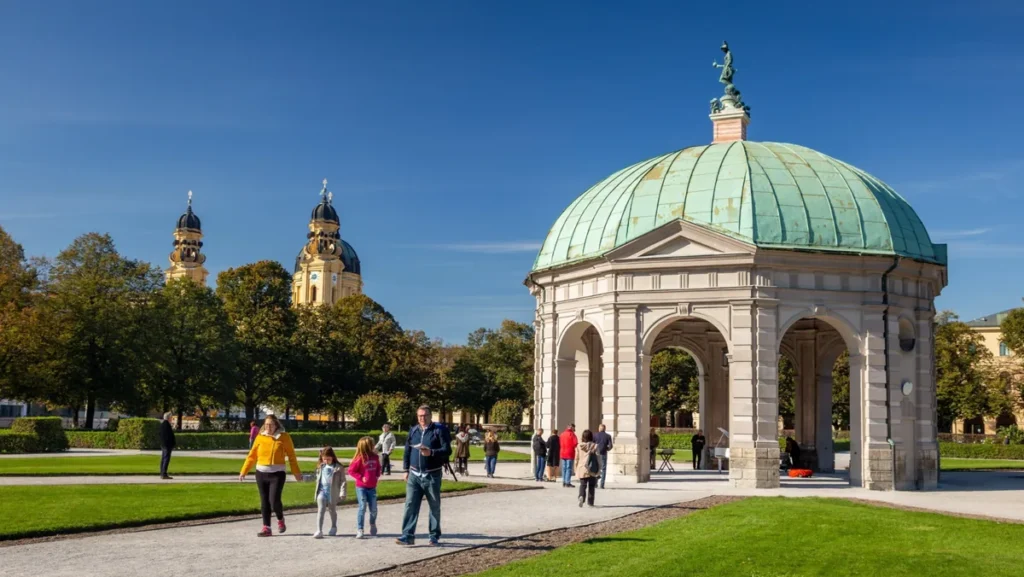 Vista do Hofgarten, um dos jardins históricos mais elegantes de Munique, na Alemanha, com destaque para o pavilhão central conhecido como Templo de Diana (Dianatempel). A construção apresenta uma cúpula de cobre esverdeada, colunas e arcos clássicos, formando um dos cenários mais característicos do jardim. Ao fundo, aparecem as torres da Theatinerkirche, igreja barroca localizada próxima à Odeonsplatz. Criado no século XVII como jardim da residência dos governantes da Baviera, o Hofgarten é atualmente um espaço público que combina arquitetura histórica, áreas verdes e caminhos tranquilos. Sua localização central faz dele uma parada popular durante os passeios pelo centro histórico de Munique.