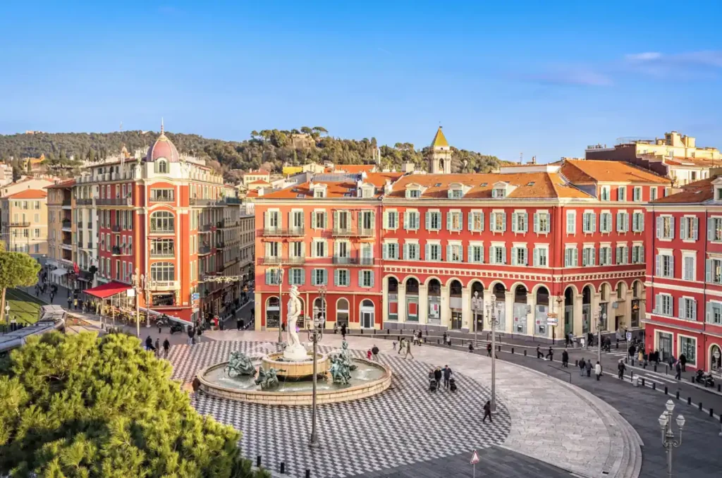 Place Masséna, uma das principais praças e atrações turísticas de Nice, na Côte d’Azur, França. A praça destaca-se por sua arquitetura neoclássica de fachadas avermelhadas, piso quadriculado preto e branco e pela Fontaine du Soleil, decorada com uma estátua de Apolo. É um dos pontos centrais para explorar o centro histórico e as atrações de Nice.