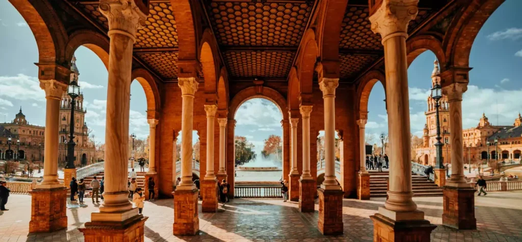 Perspectiva arquitetônica das galerias cobertas da Plaza de España, em Sevilha, destacando a beleza de suas colunas de mármore, arcos de tijolos avermelhados e tetos decorados com padrões geométricos. A composição da fotografia cria um efeito de profundidade através das arcadas, que enquadram a fonte central da praça e as pontes ornamentadas ao fundo. Construída no início do século XX, a Plaza de España é um dos maiores símbolos arquitetônicos da cidade e um exemplo marcante da combinação de estilos tradicionais espanhóis. O local atrai visitantes por seus detalhes artísticos, azulejos decorativos e espaços ideais para caminhadas e fotografias.